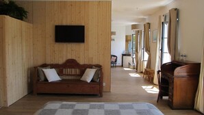 A room with a wooden wall, a sofa with cushions, a flat-screen TV, a desk with a chair, and a window with curtains. - A l'Ecole Buissonnière (Honfleur)