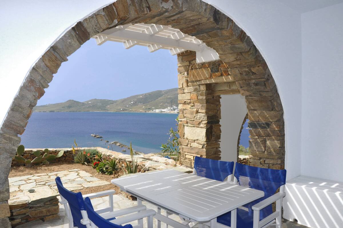 A stone archway leading to a terrace with a white table and blue chairs, overlooking a coastal landscape.
