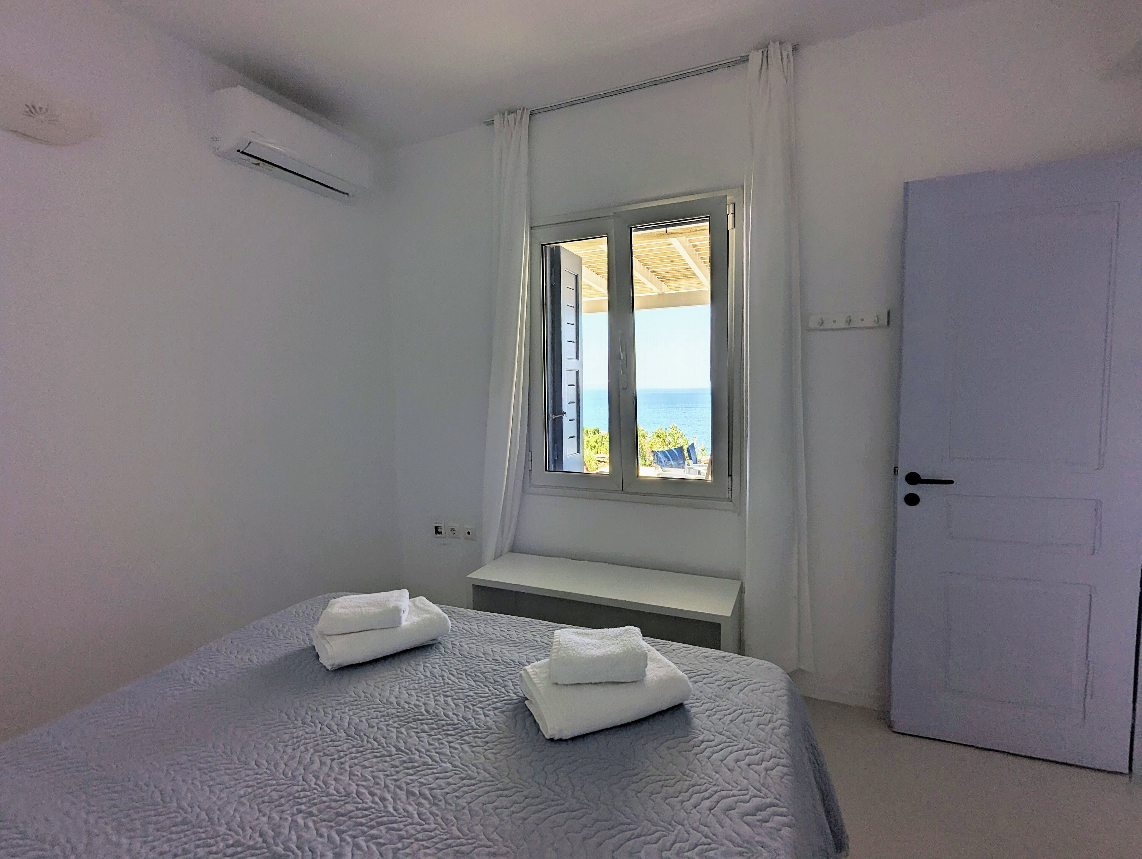 A bedroom with a bed, two towels, a window with a view, an air conditioning unit, and a door.