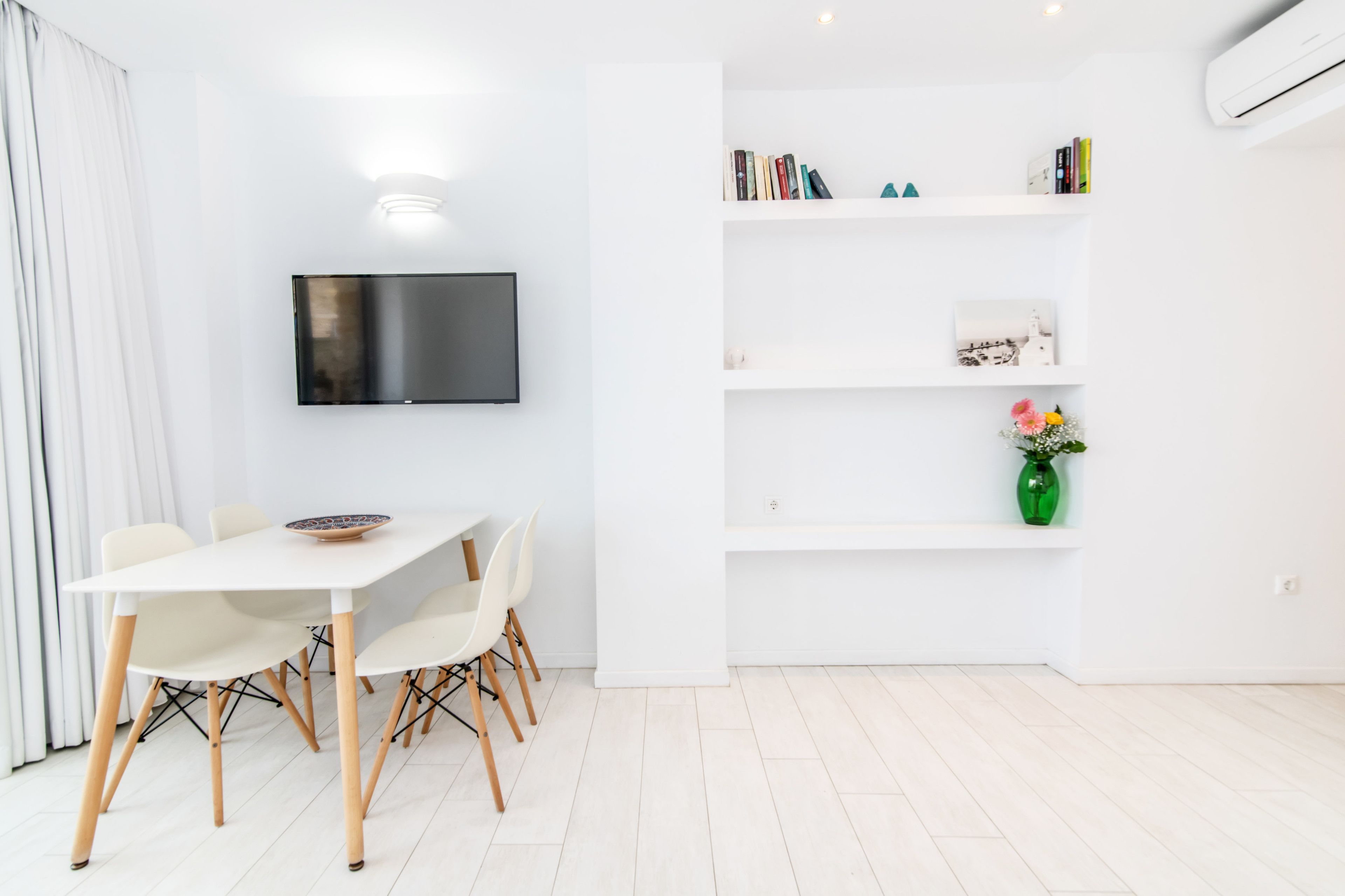 A modern dining area with a white table and chairs, a flat-screen TV mounted on the wall, and a bookshelf with books and decorative items.