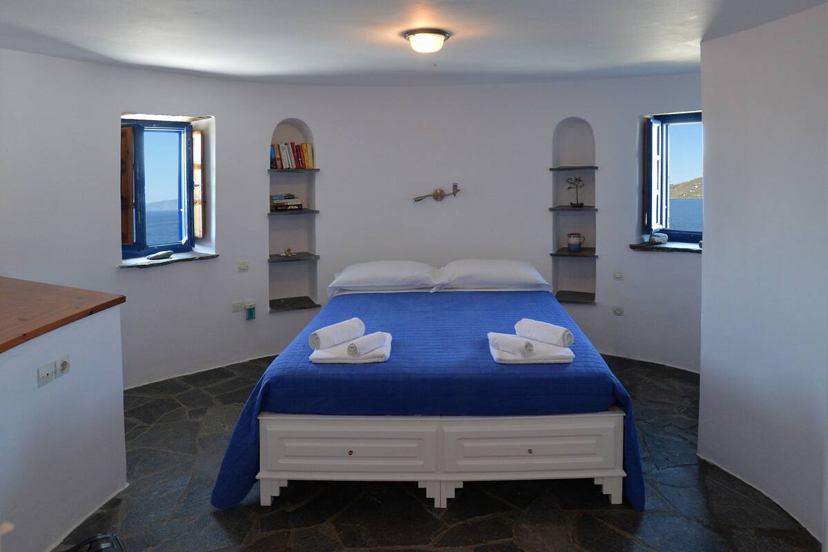 A bedroom with a bed, two windows, a shelf with books, and a stone floor.