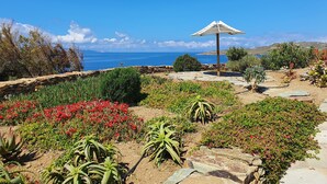 A garden with a stone pathway, red flowers, and a white umbrella. - Stavros Bay (Tinos)