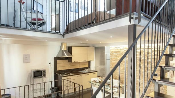 Una cocina moderna con una isla central, una zona de comedor con mesa de vidrio y una escalera que conduce a un nivel superior.