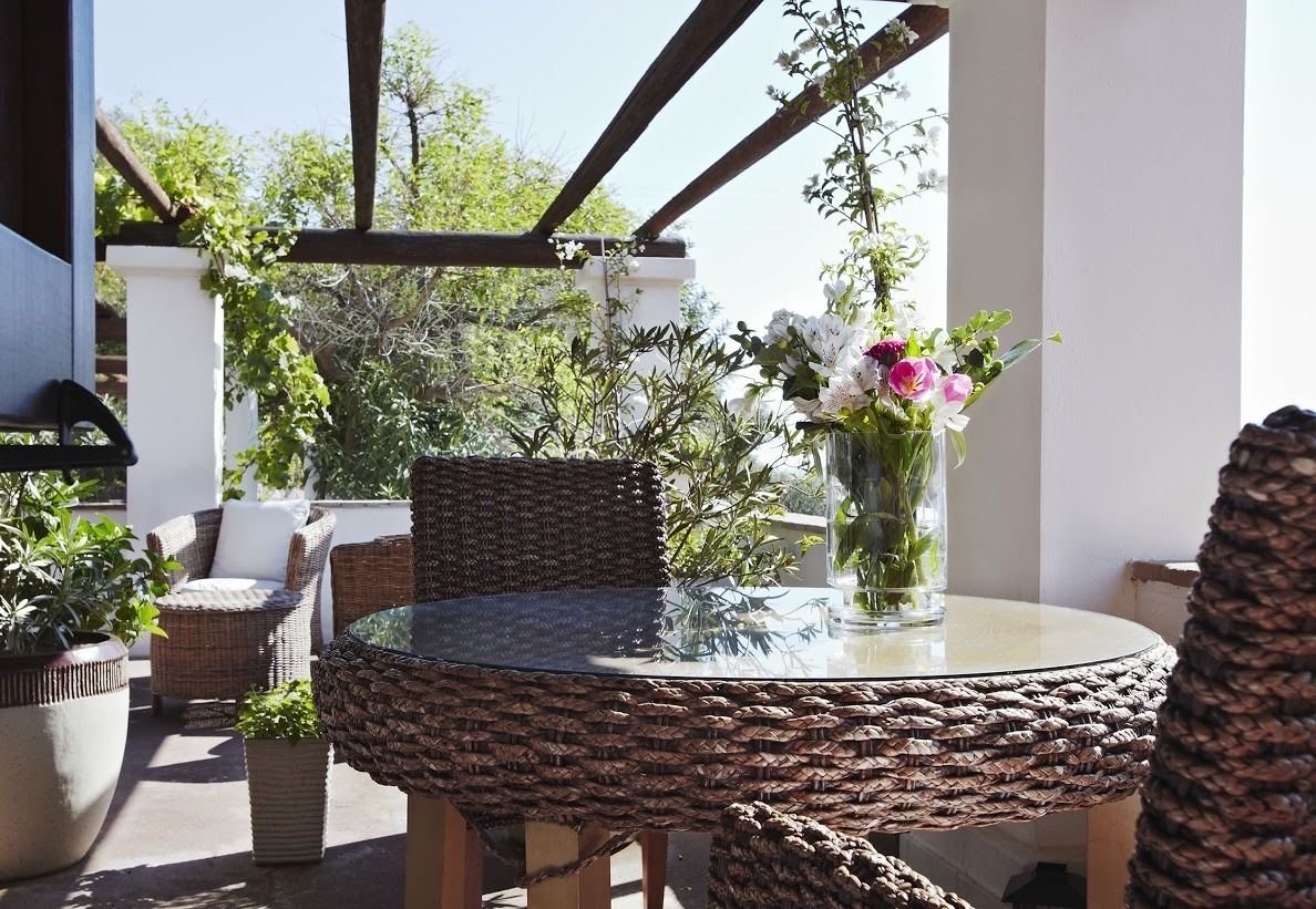 Een terras buiten met rieten meubels, een glazen tafel en een vaas met bloemen.