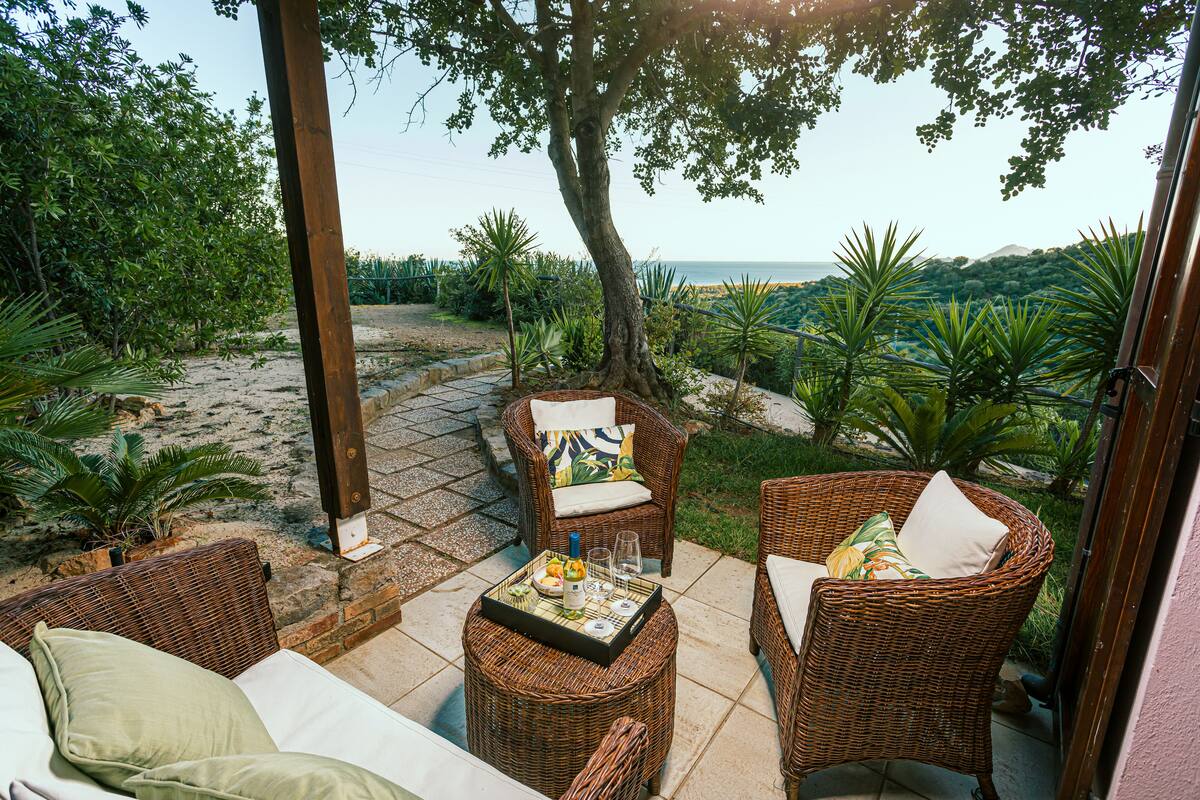 A covered outdoor seating area with wicker furniture and a table set with a bottle, glasses, and a tray.