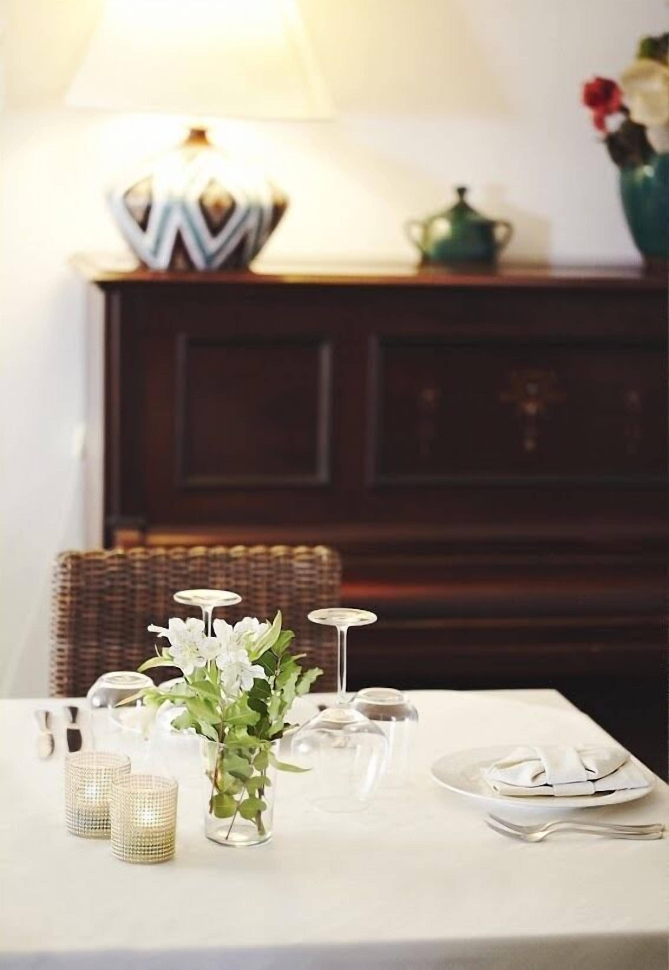 A dining table set with white tableware, a vase with white flowers, and a lamp.