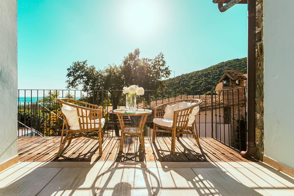 A balcony with wicker furniture and a table with a vase of flowers, overlooking a scenic view of a coastal area.