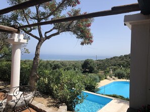 A pool area with a view of the sea, surrounded by greenery and a pergola structure.
