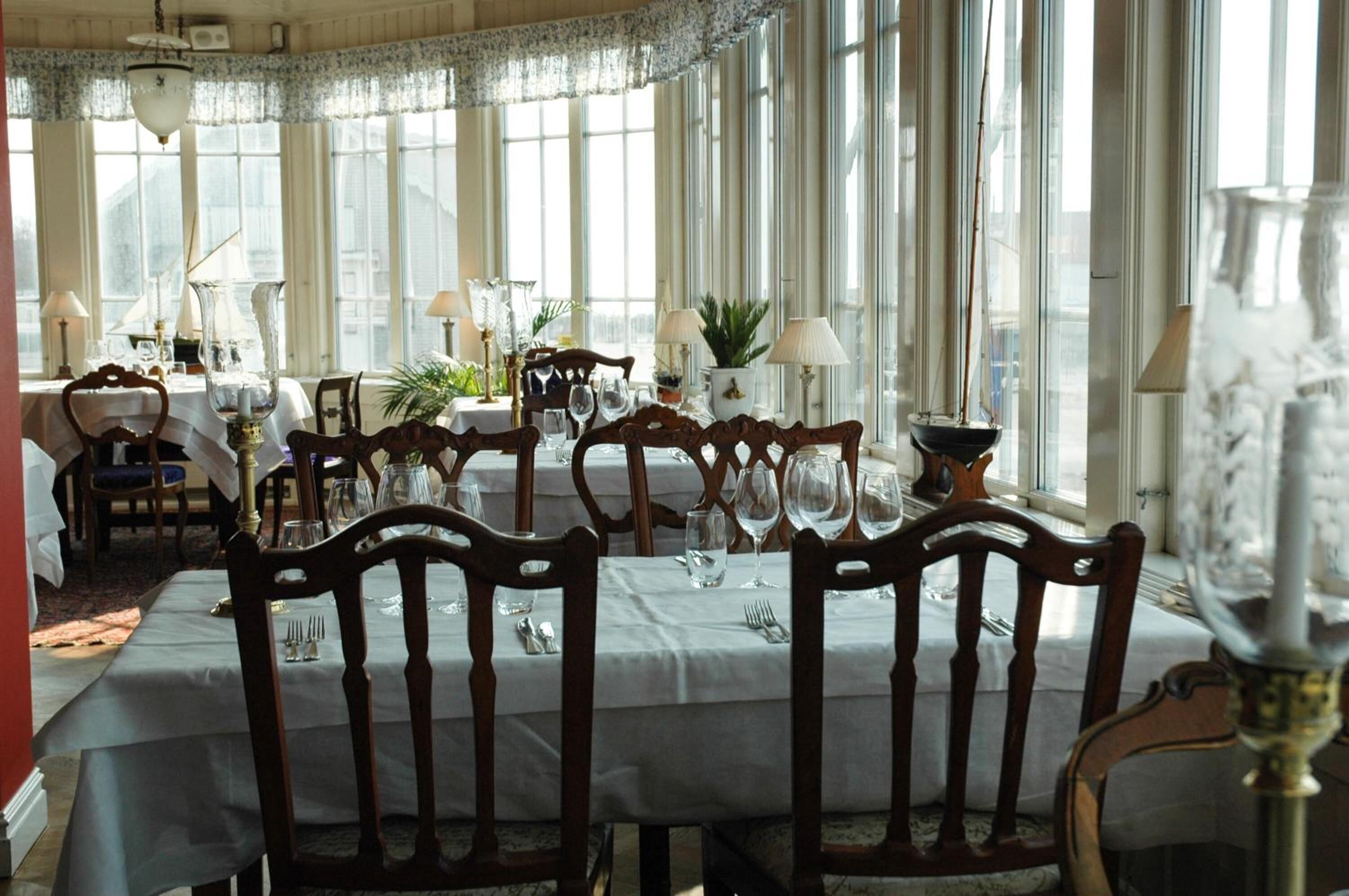 Un comedor con mesas dispuestas para una comida, ventanas amplias y decoración clásica.