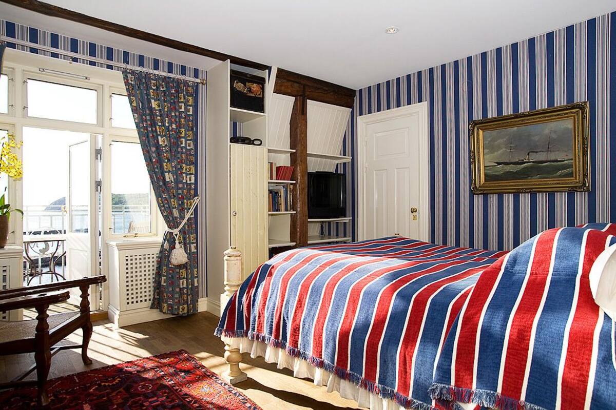 Um quarto com uma cama, janela com cortinas, estante de livros e um quadro na parede.