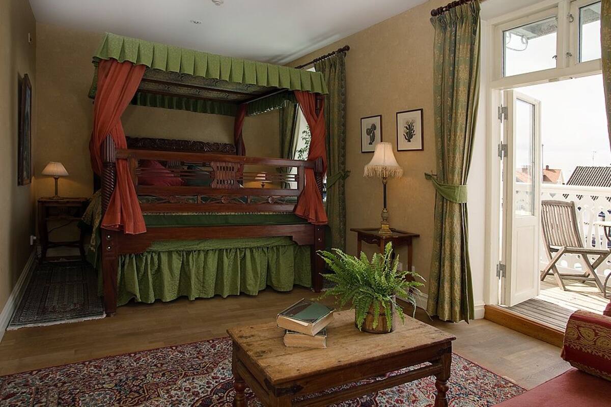 Quarto com cama de dossel, mesa de centro de madeira com livros, planta em vaso e vista para uma varanda com móveis de exterior.
