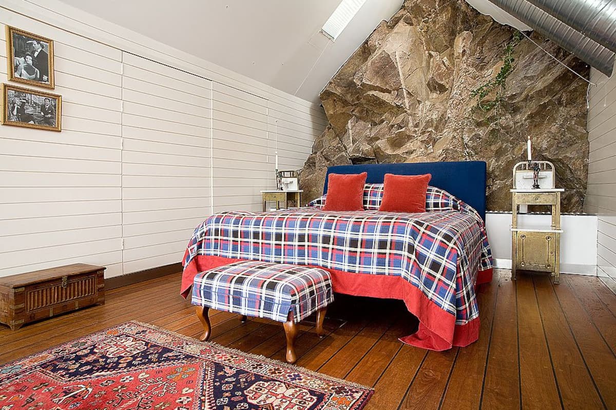 Quarto com teto inclinado, cama com colcha xadrez, um banquinho, um tapete e parede de pedra.