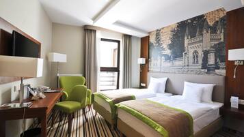 Quarto de hotel com duas camas, uma escrivaninha com computador, uma cadeira verde e uma janela grande.