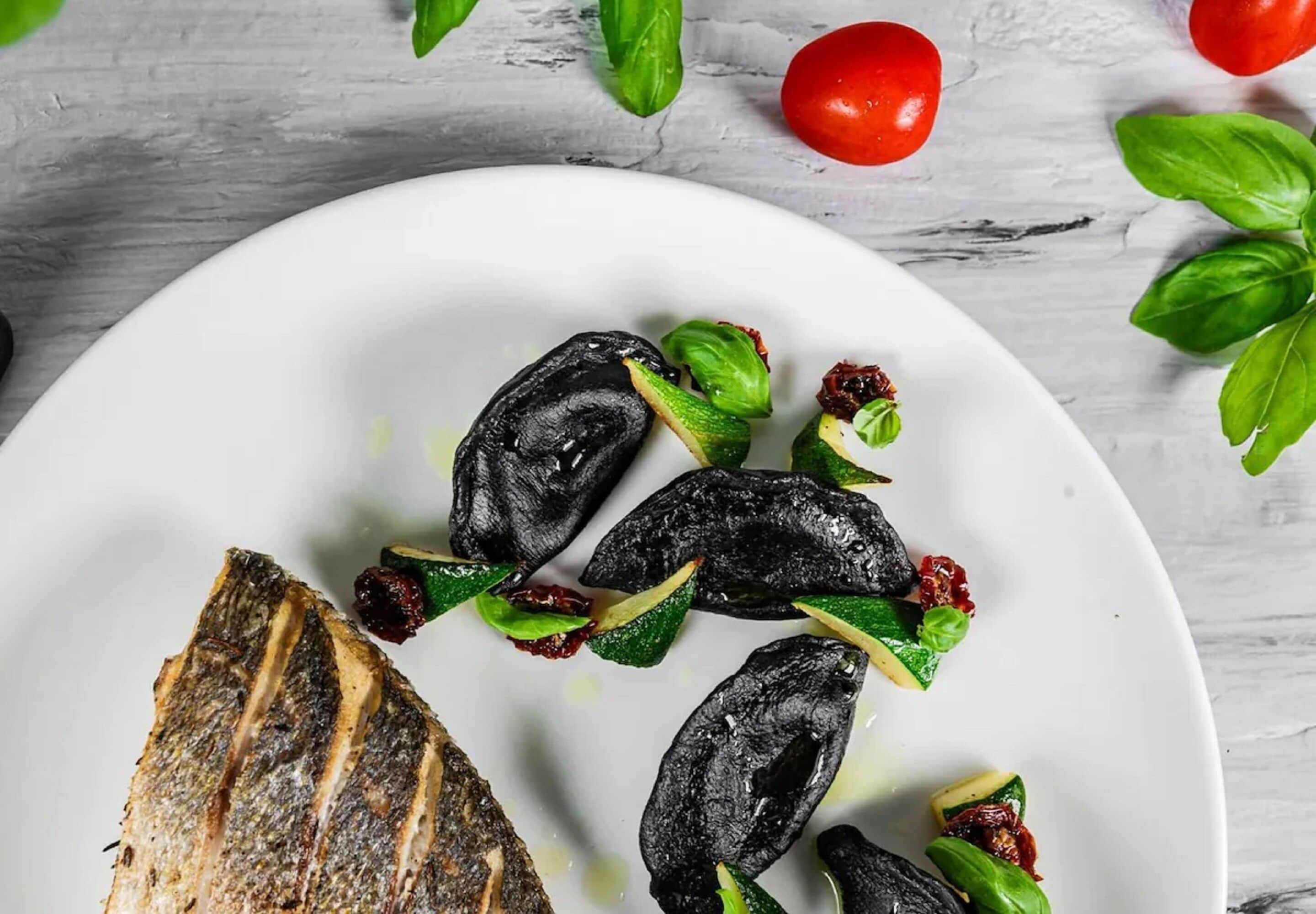 A plate with grilled fish, black olives, and green leaves on a white plate.