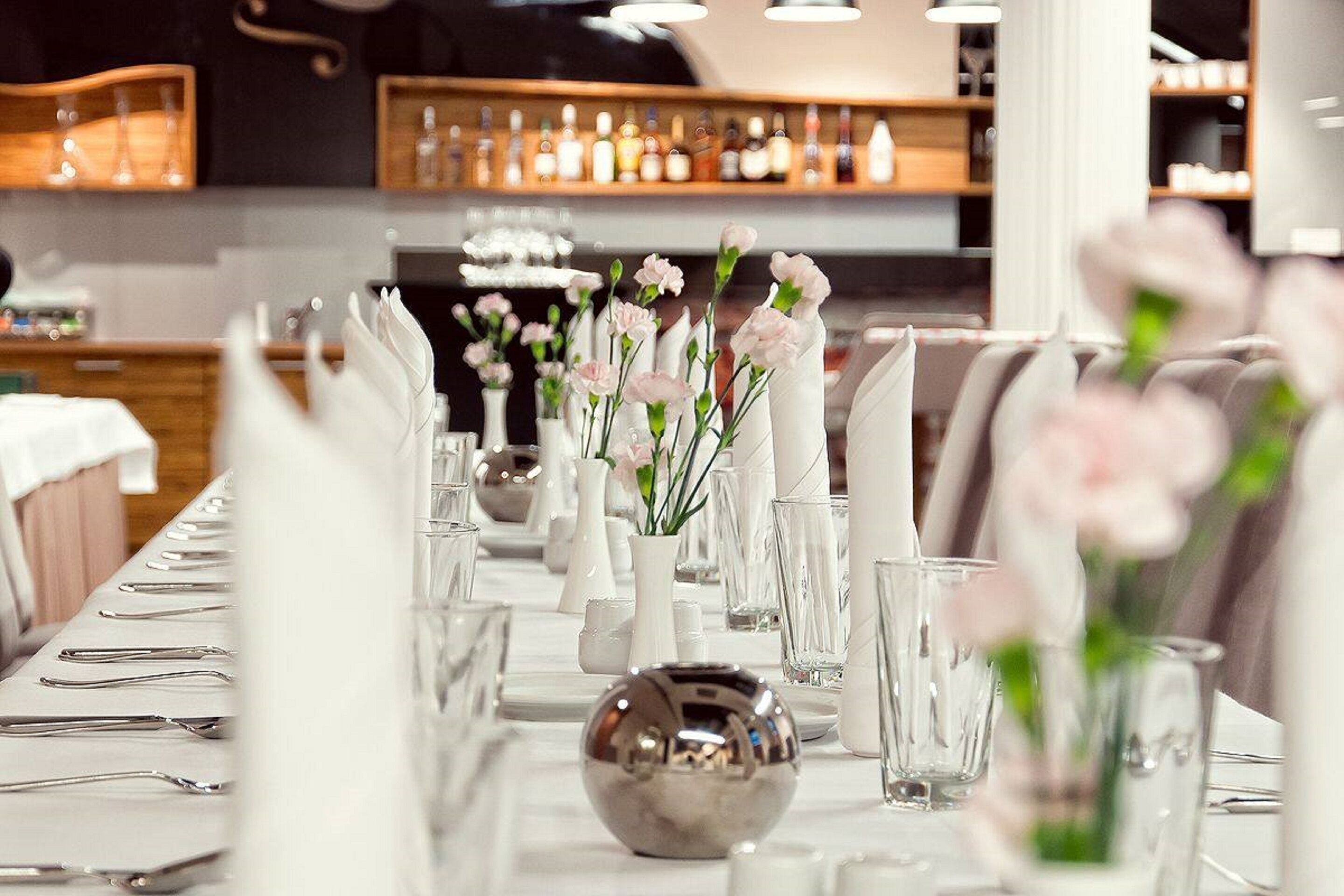Een lange eettafel, gedekt met witte servetten, bestek en glazen, met bloemstukken in het midden en een bar op de achtergrond.