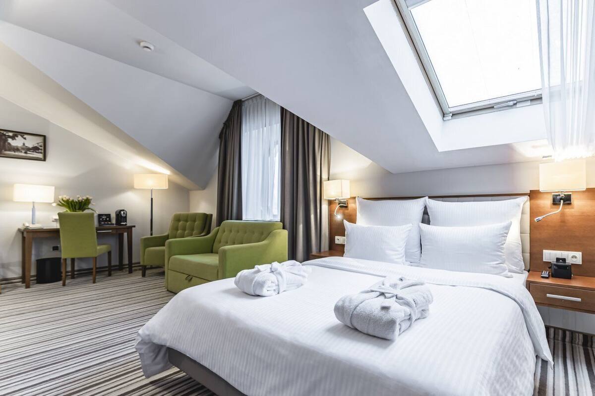 A hotel room with a large bed, two green armchairs, a desk with a lamp, and a view of the sky through a skylight.