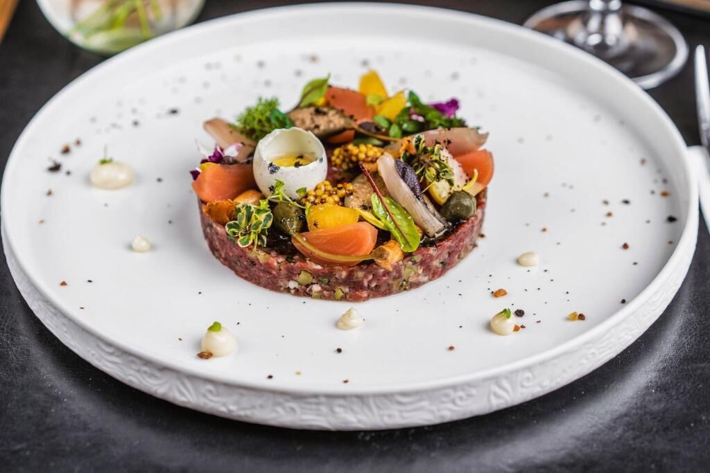Un plato blanco con tártara de carne, adornado con diferentes verduras y una yema de huevo cruda.