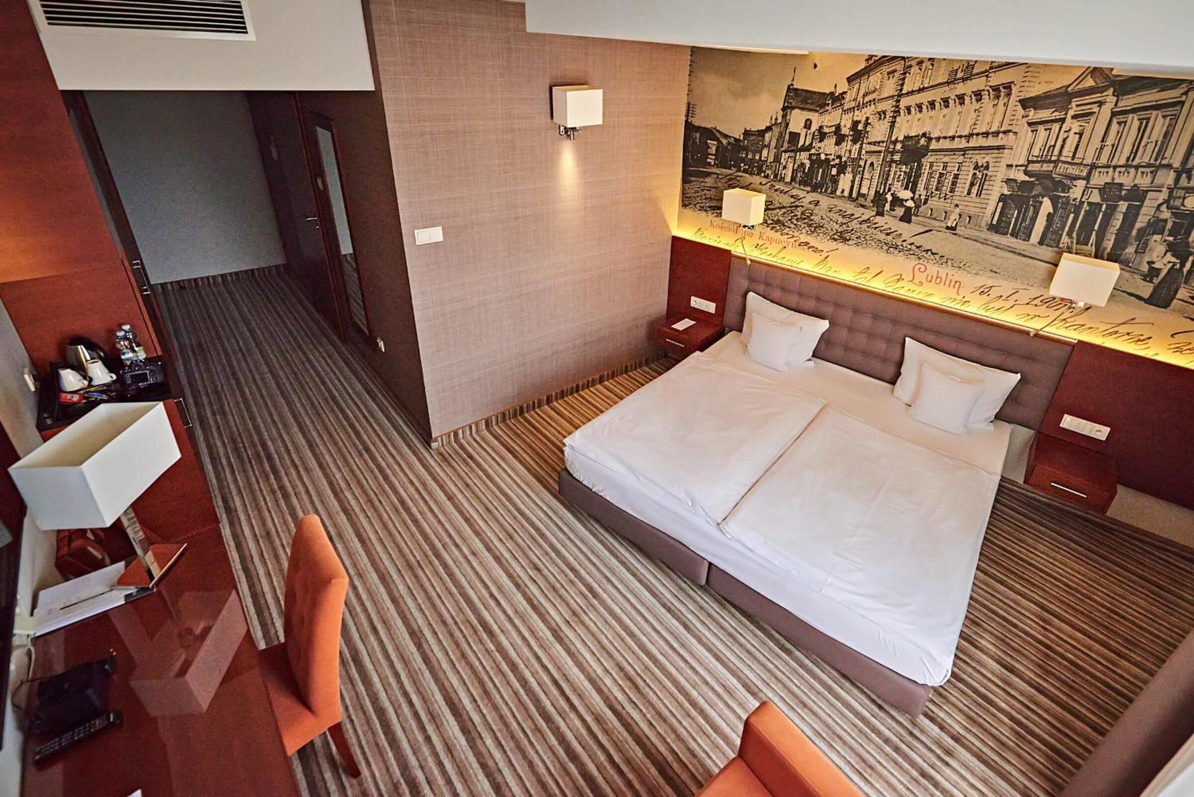 A hotel room with a large bed, a desk with a chair, a nightstand, and a wall-mounted lamp.
