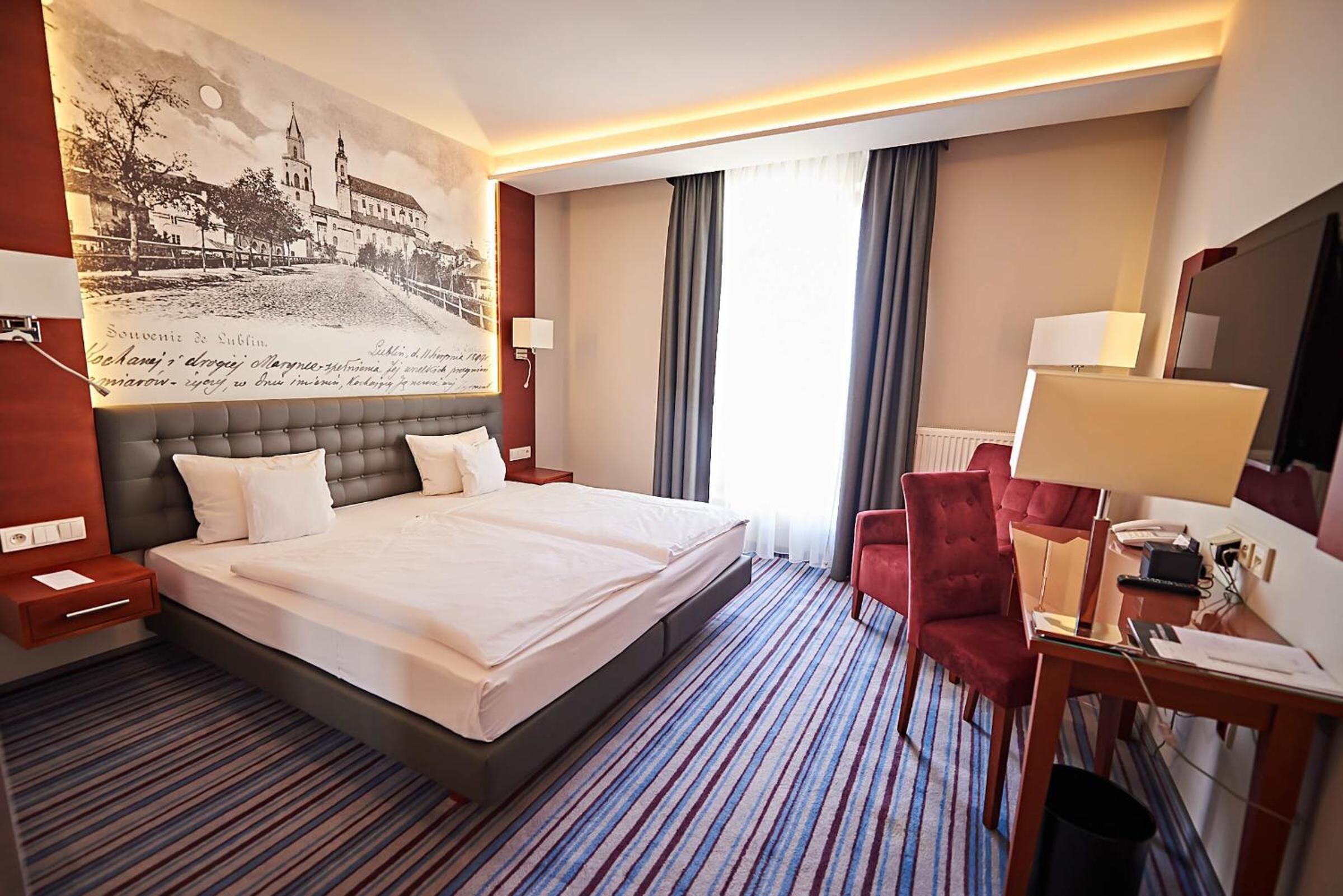 Een hotelkamer met een groot bed, een bureau met stoel, een wandtelevisie en een groot raam met gordijnen.