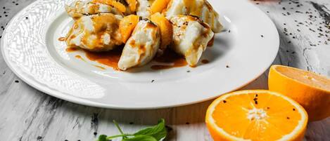 A plate of dumplings with a sauce, accompanied by whole and halved oranges and scattered peppercorns.