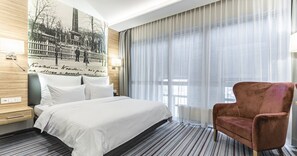 Une chambre d’hôtel avec un grand lit, un fauteuil rouge, un téléviseur fixé au mur et une fenêtre ornée de rideaux légers.