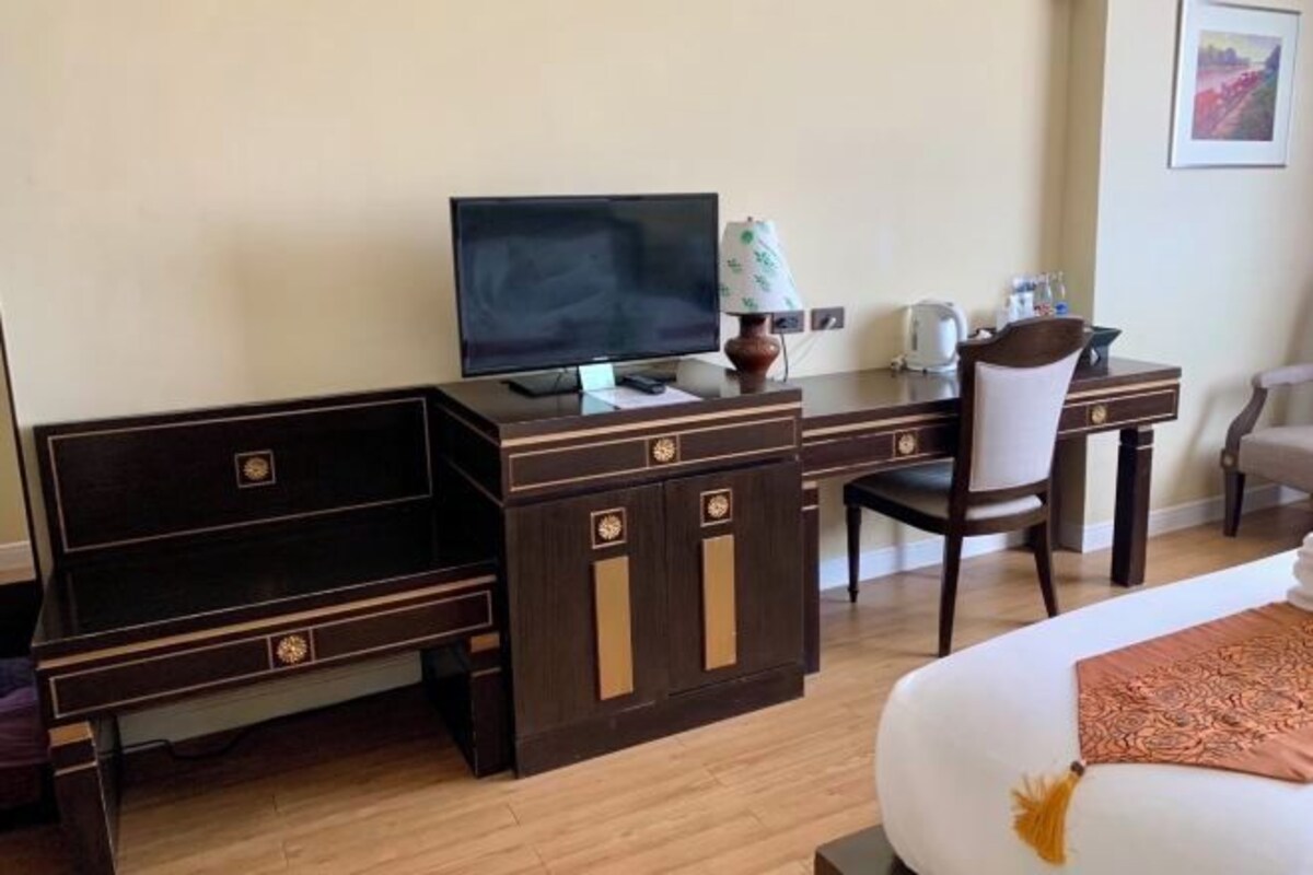 A hotel room with a bed, a desk with a television, a chair, and a small table with a lamp.
