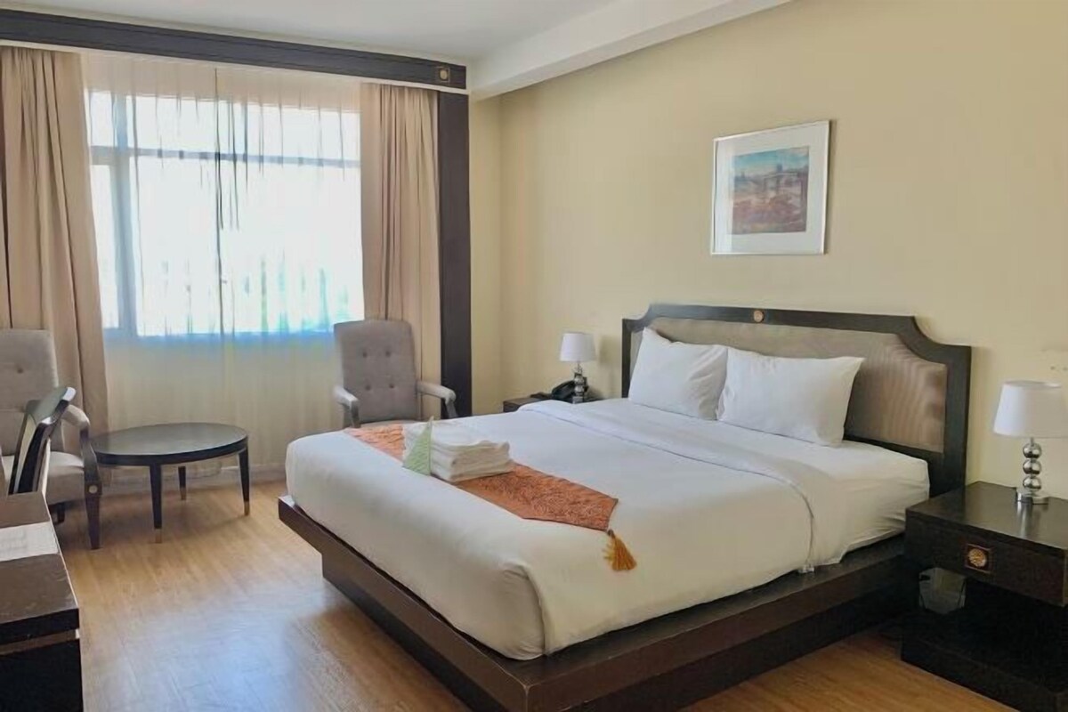 A hotel room with a large bed, two bedside tables, a chair, a desk, and a window with curtains.