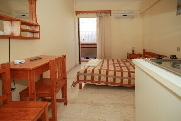 A compact hotel room with a wooden desk, a bed with a checkered bedspread, a small bedside table, and a window with curtains. - Astir Rooms (Kefalonia)