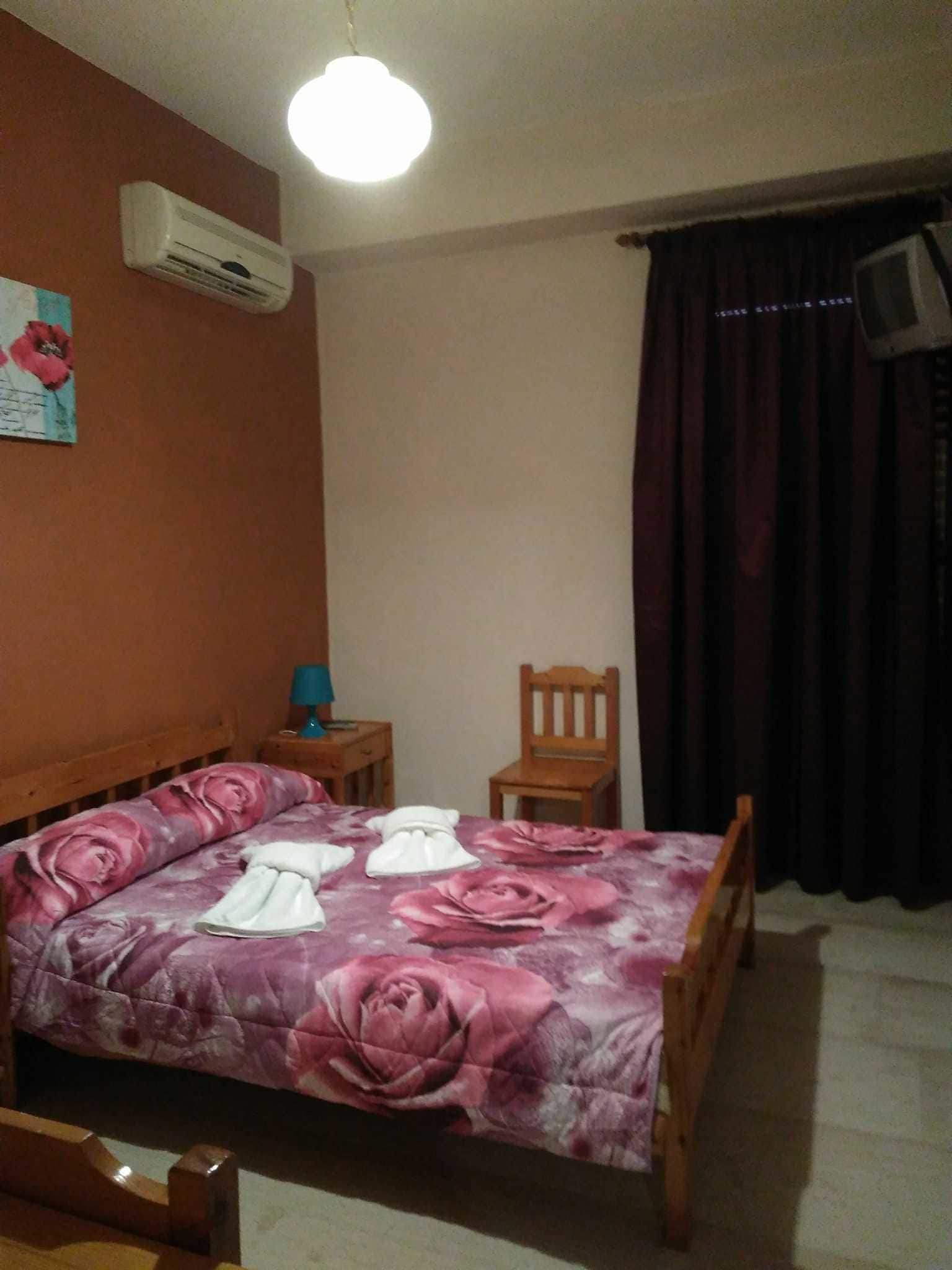 Un dormitorio con una cama, una silla de madera, una mesita de noche, un aire acondicionado y una televisión.