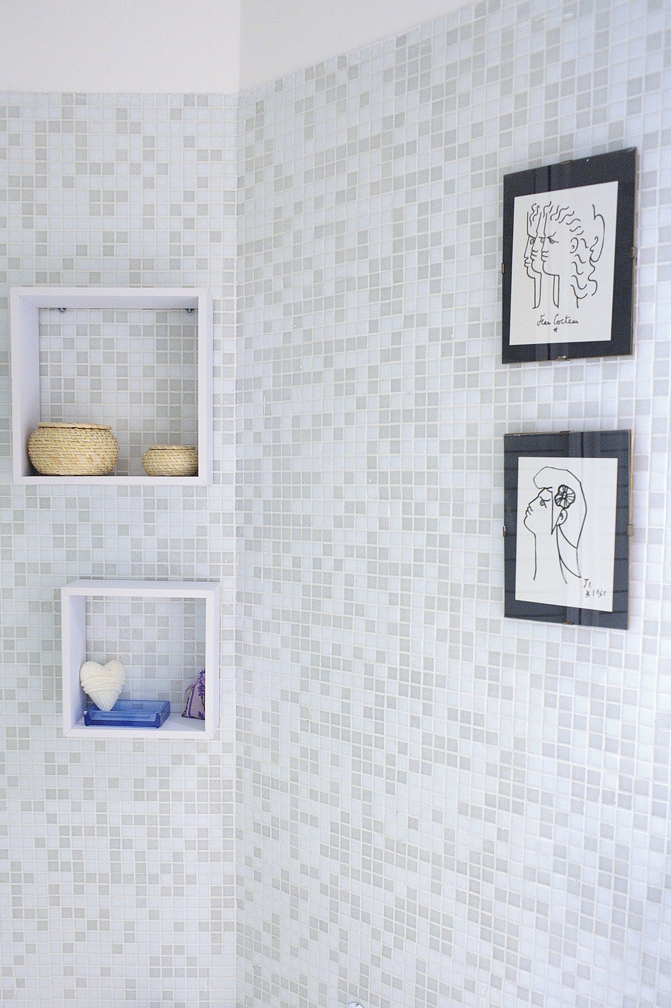 Een badkamer met een betegelde muur, twee ingelijste kunstwerken en twee planken met manden en een zeepbakje.