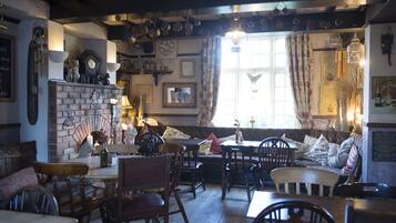 Un pub acogedor con mobiliario de madera, chimenea de ladrillo y diversas decoraciones, incluyendo un reloj y luces colgantes.