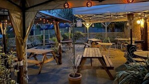 Un espace extérieur aménagé en terrasse, avec des tables et des bancs en bois, le tout recouvert d’une pergola illuminée de guirlandes lumineuses.