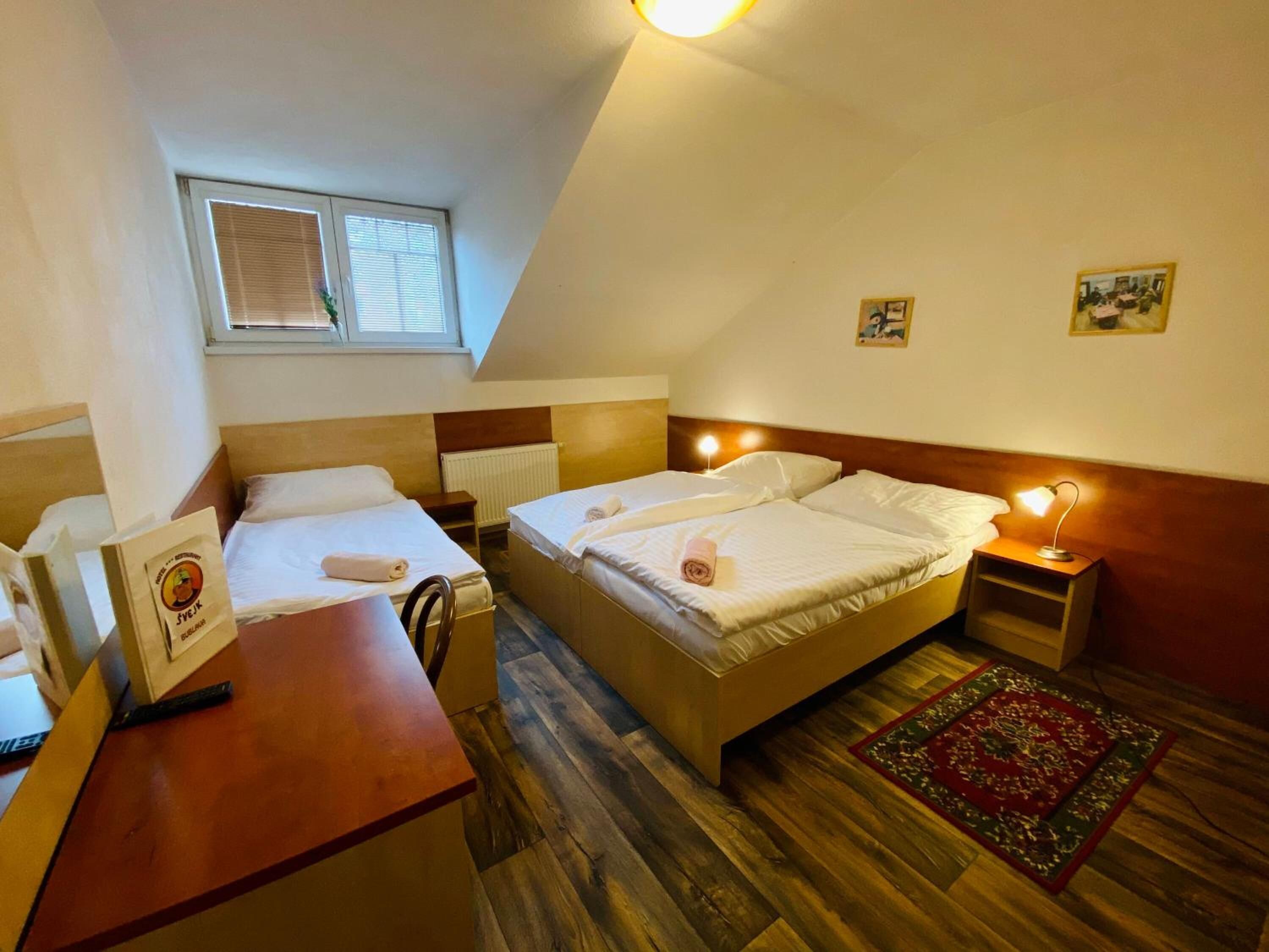 Ein Hotelzimmer mit zwei Betten, einem Holztisch, einem kleinen Tisch, einem Fenster mit Jalousien, einer Heizung und einem roten Teppich mit Muster.