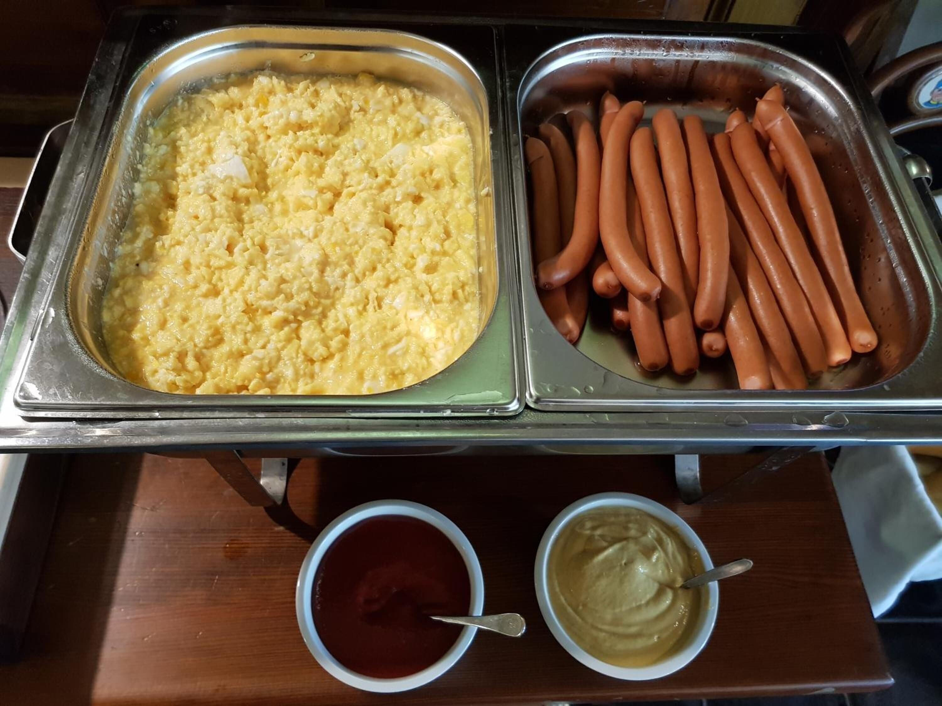 Una mesa de buffet con huevos revueltos, perros calientes, kétchup y mostaza.