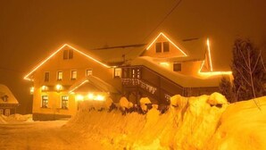A snow-covered house with a steep roof and multiple windows, illuminated by warm interior lighting. - Hotel Restaurant Švejk (Bublava)