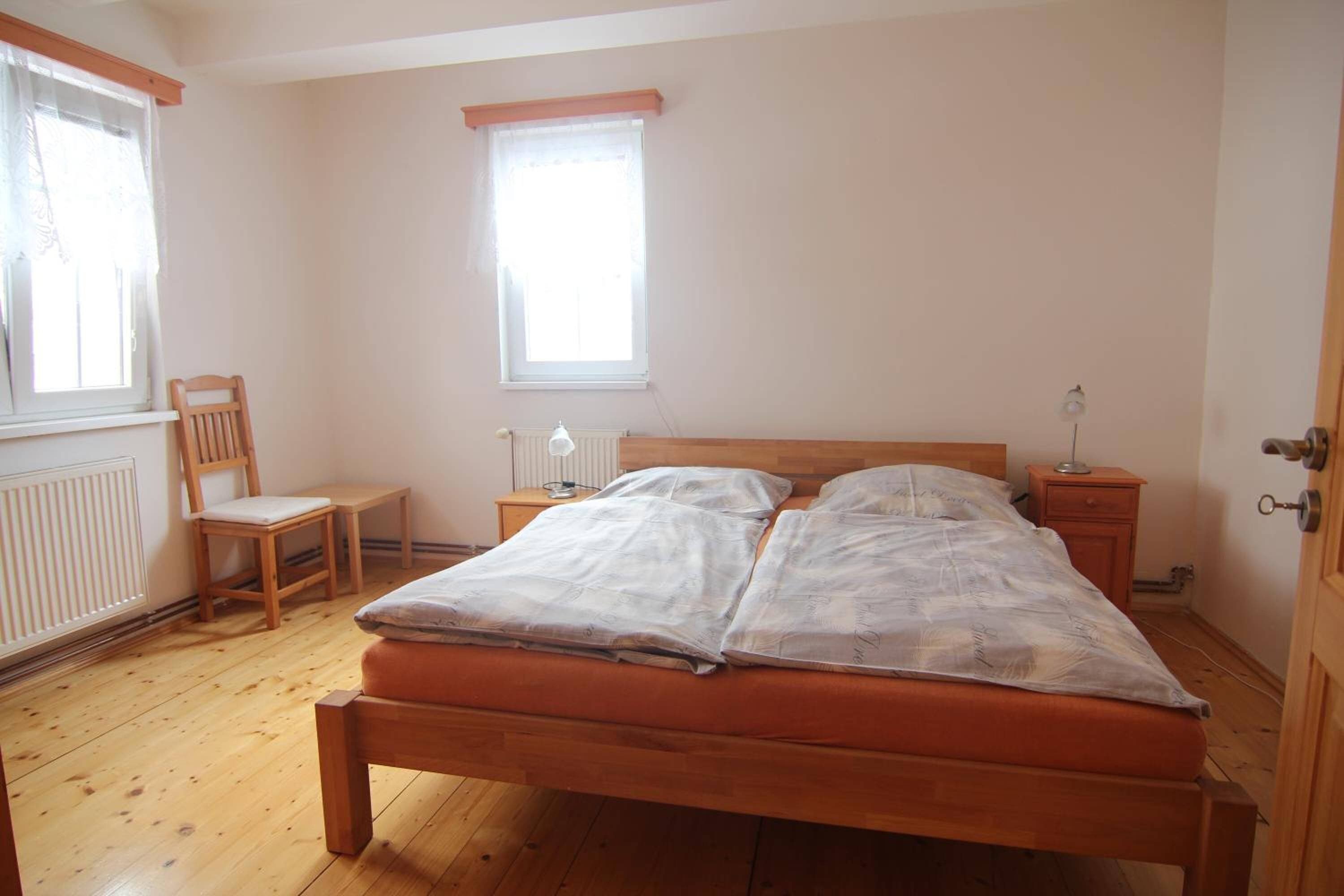 A bedroom with a wooden bed, a chair, a small table, a nightstand, a lamp, and a window.