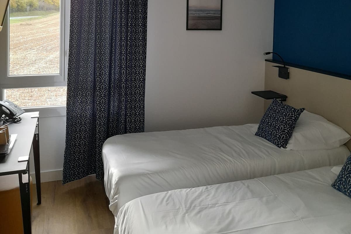 Habitación de hotel con dos camas, un escritorio con teléfono, una ventana con cortinas, un cuadro enmarcado en la pared y una pared de acento azul.