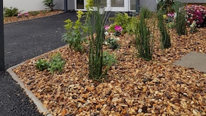 A modern building with a glass door entrance, surrounded by a garden with various plants and flowers. - Le Riccoty (Blyes)