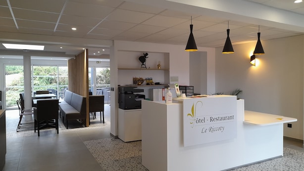 A hotel reception area with a white counter, black pendant lights, and a view of the outdoor seating area. - Le Riccoty (Blyes)