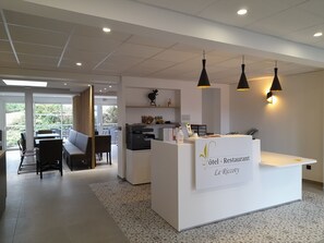 A hotel reception area with a white counter, black pendant lights, and a view of the outdoor seating area.