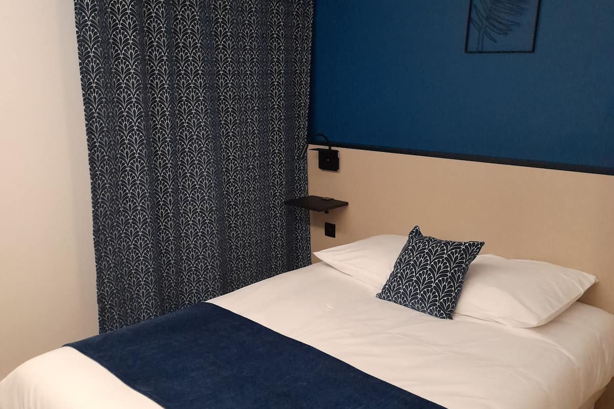 Un dormitorio con una cama, una pared azul, una obra de arte con motivos de hojas y una cortina estampada.