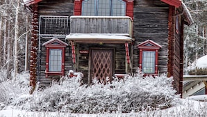 Una cabaña de madera con techo rojo y dos ventanas, rodeada de árboles cubiertos de nieve.