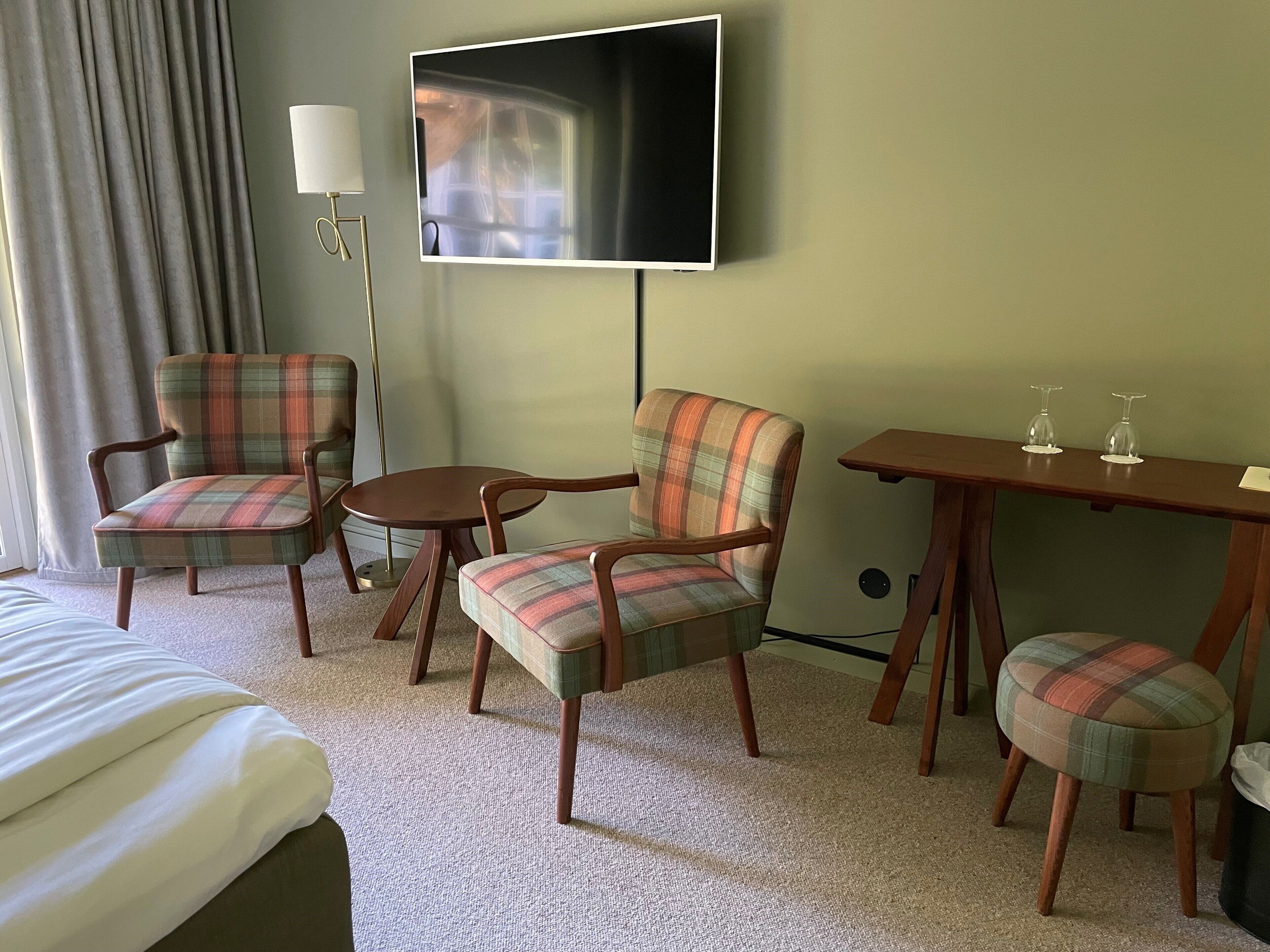 Ein Hotelzimmer mit einem Bett, zwei karierten Sesseln, einem kleinen Tisch, einem Fernseher und einer Lampe.