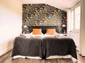 Un dormitorio con papel tapiz floral, cabecera negra, dos almohadas naranjas y dos camas tapizadas en gris.