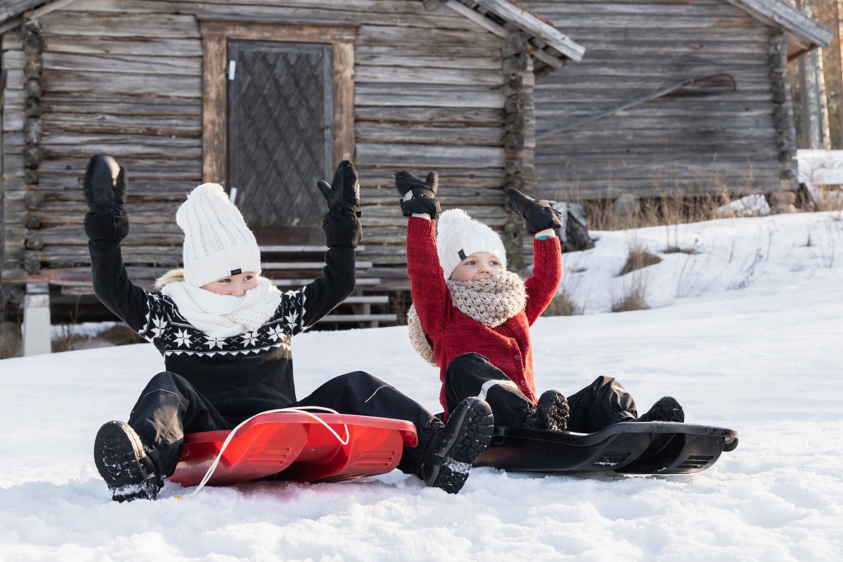 Two people sledding on snow in front of a wooden cabin.
