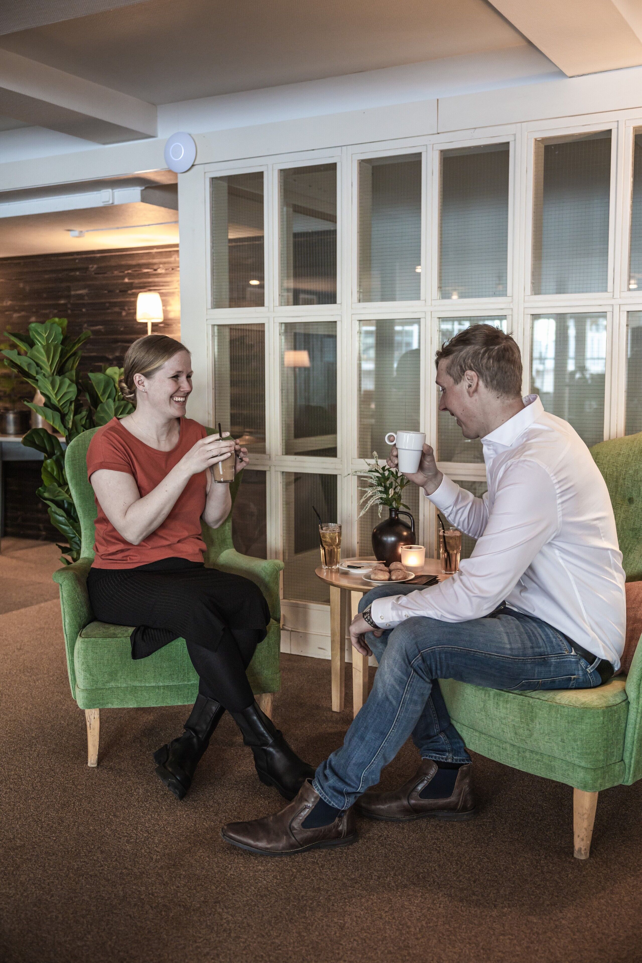Two people seated on green chairs at a small table with drinks and a candle, in a room with large windows and a plant in the background.