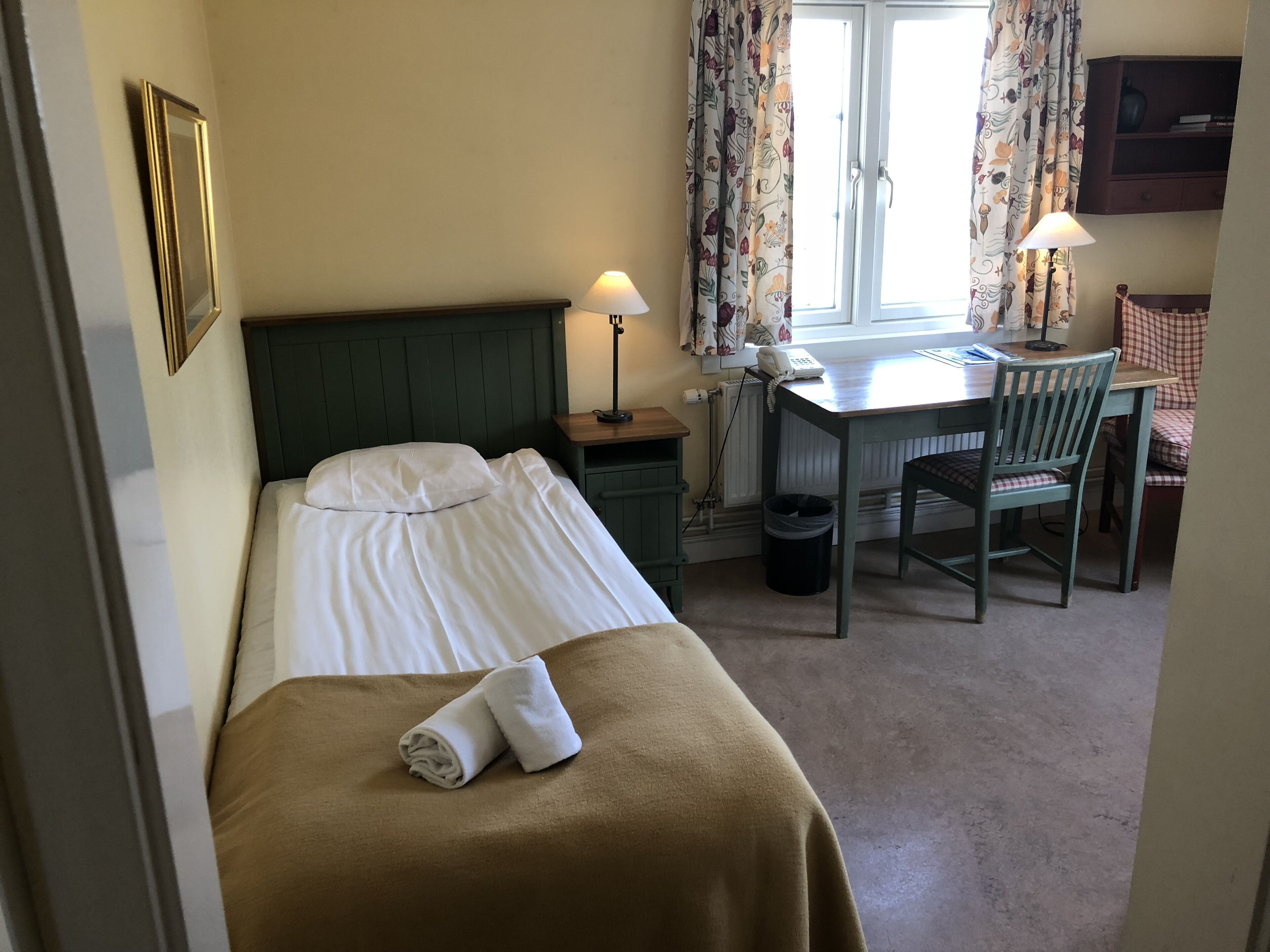 A hotel room with a bed, a desk, a chair, a lamp, and a window with curtains.