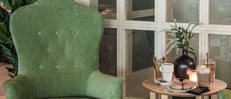 Un sillón verde, una mesita redonda pequeña con bebidas y aperitivos, y un ventanal con persianas.