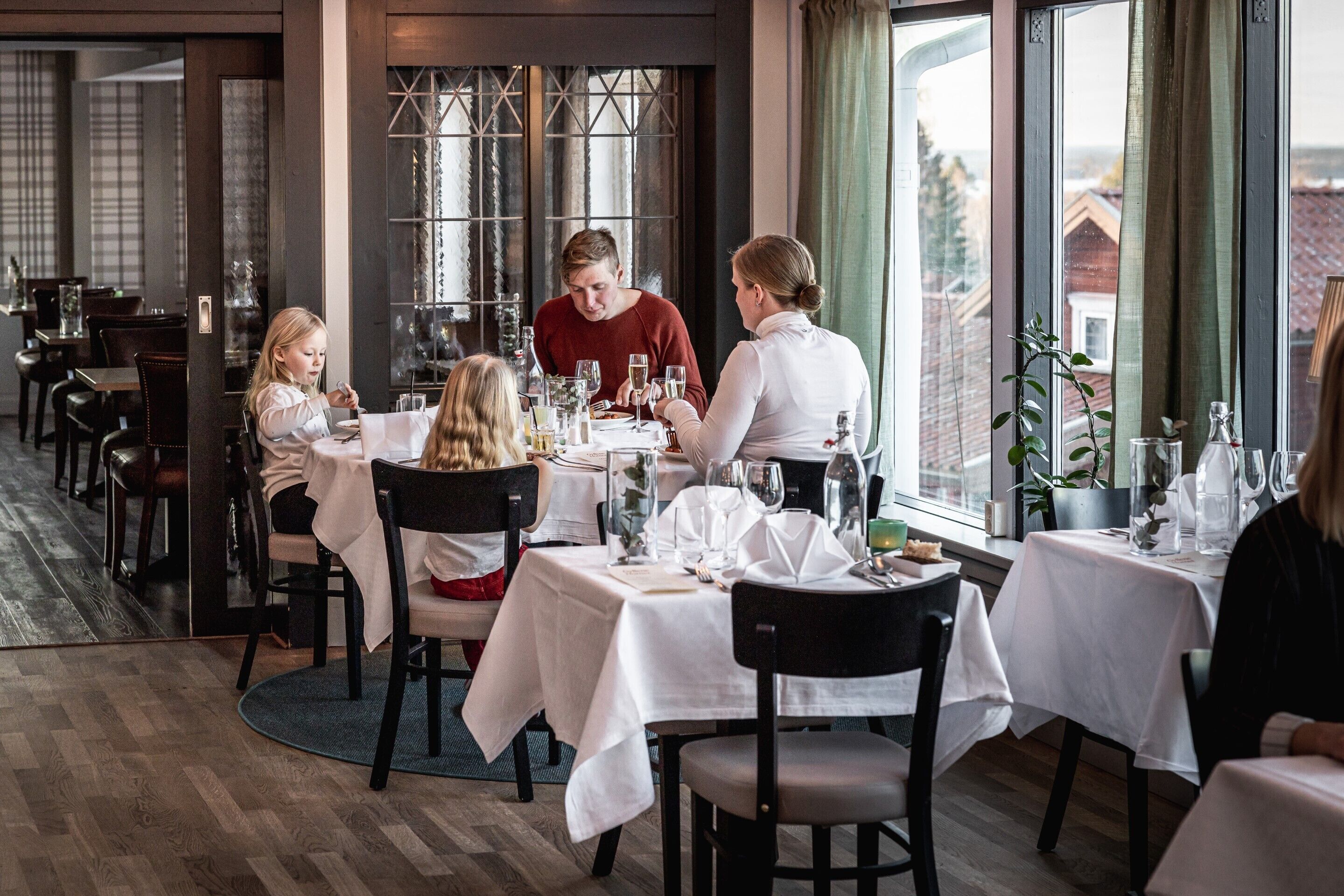 Ein Restaurant mit runden Tischen, weißen Tischdecken und Blick auf Gebäude durch große Fenster.