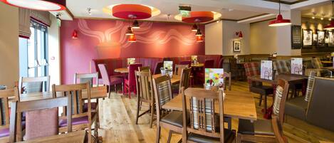 A restaurant interior with wooden tables and chairs, red pendant lighting, and a wavy wall design.